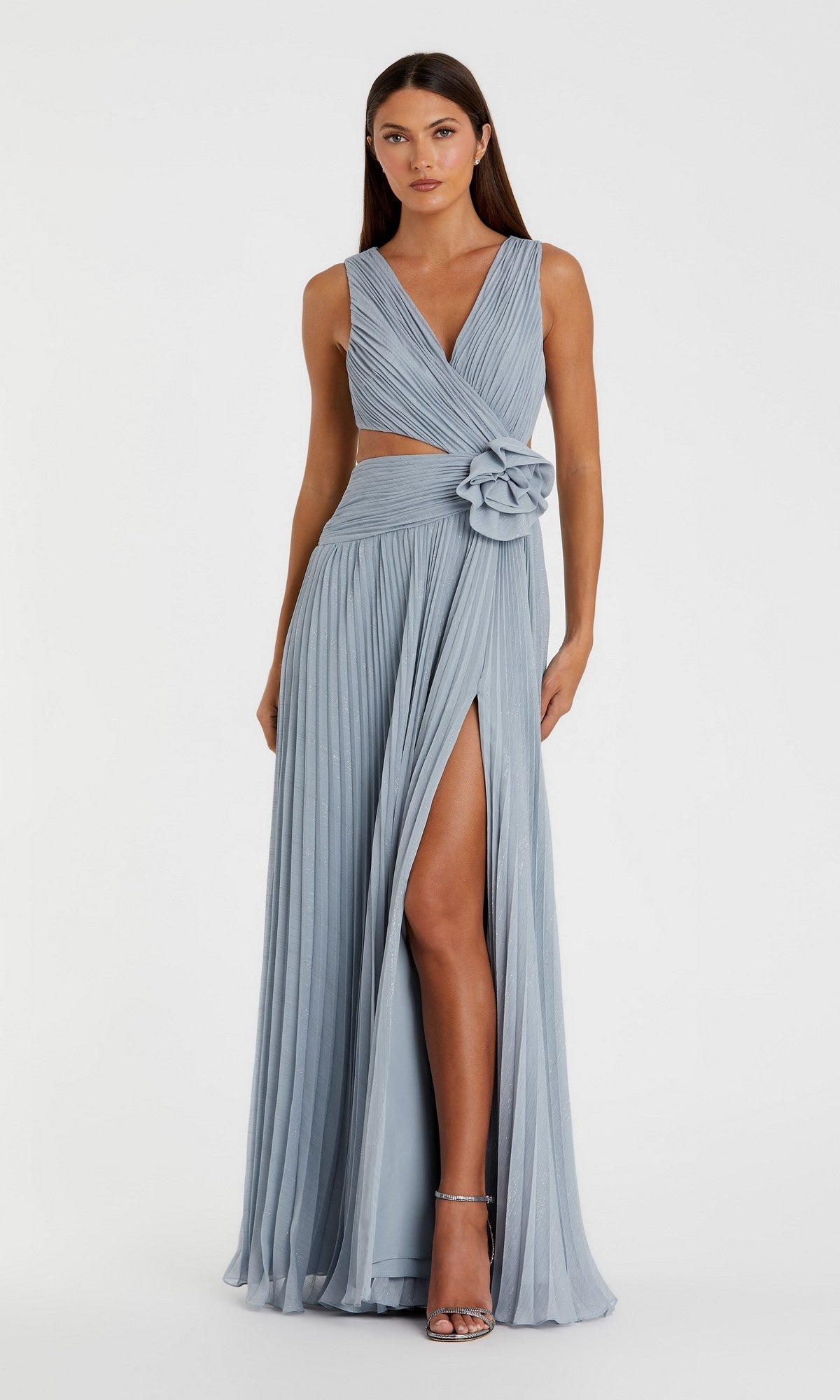 A woman stands against a plain background in the Lilac Long Pleated Formal Dress: Mac Duggal 49938, featuring a sleeveless design, high slit, side cutout, and a large flower at the waist.